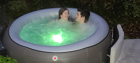 Paar entspannt in einem runden Canadian Spa Company Whirlpool im Garten bei Abenddämmerung, beleuchtet durch grünes LED-Licht im Wasser, das eine stimmungsvolle Atmosphäre schafft.